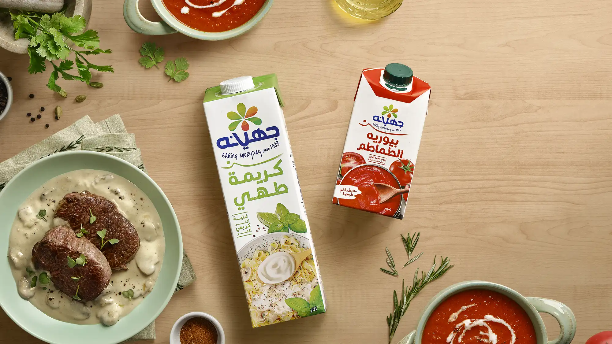 Juhayna Cooking Cream and Tomato Puree cartons in an overhead flat-lay styled with a creamy beef medallion dish, tomato soup, fresh herbs, and spices on a natural wood surface
