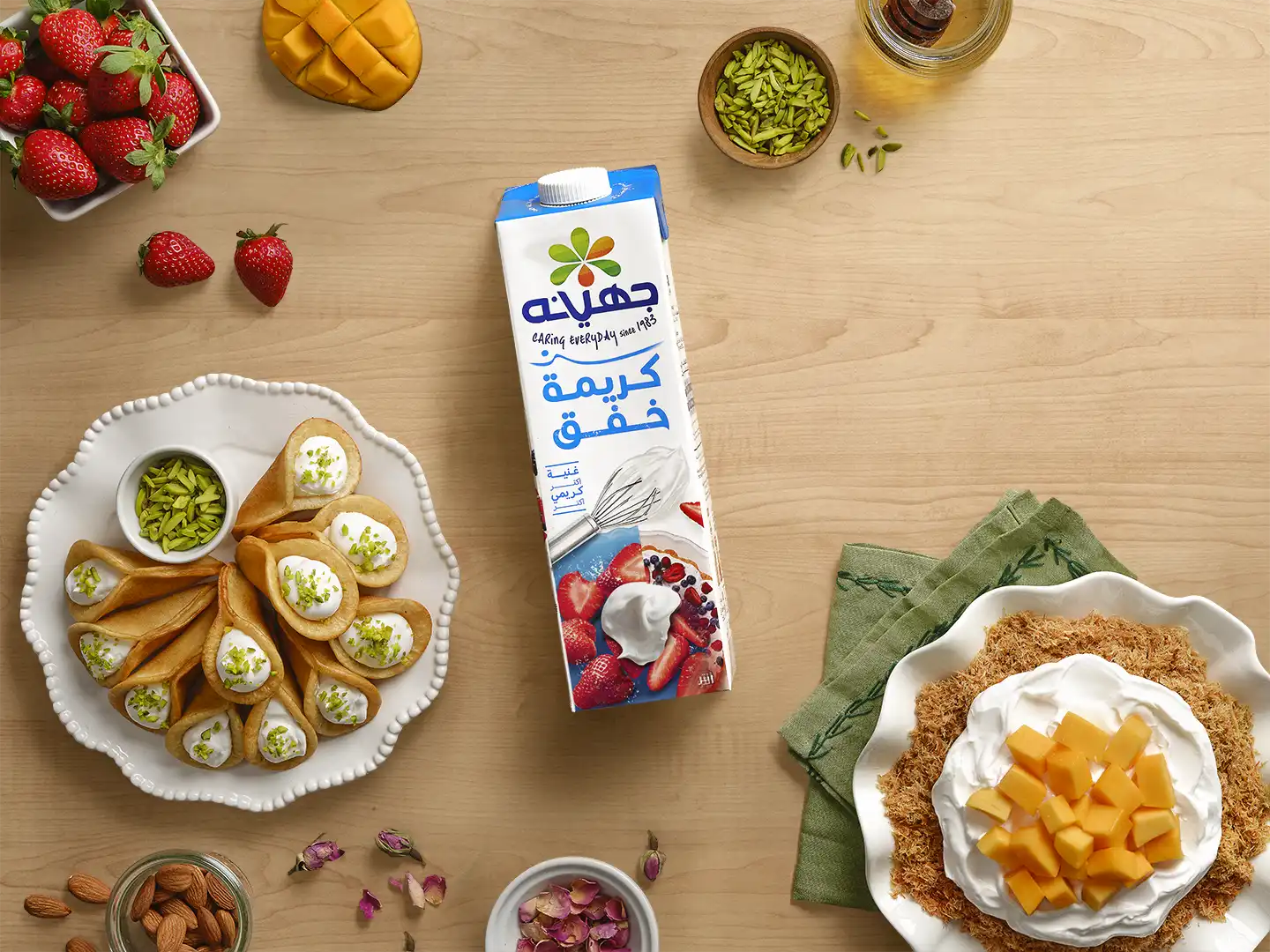 Juhayna Whipping Cream 1-litre carton in a flat-lay overhead shot surrounded by strawberries, mango, pistachios, rose petals, and two cream desserts including qatayef and a mango cream dish