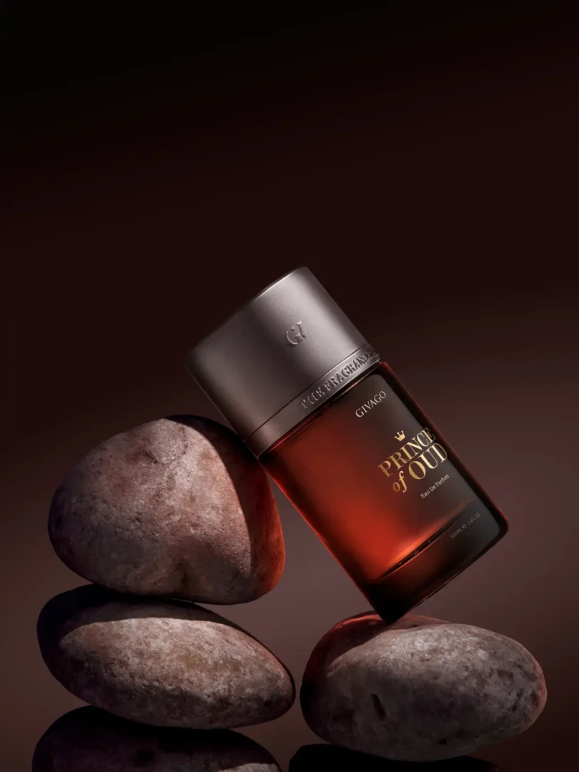 svg+xml;charset=utf - givago the one perfume photography Givago Prince of Oud perfume photography: amber bottle balanced on stacked river stones against a dark brown backdrop